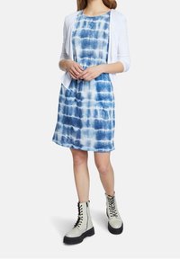 Blaue Batikkleid mit Rundhalsausschnitt und kurzen Ärmeln, kombiniert mit einem weißen offenen Cardigan und grauen Schnürstiefeln mit schwarzen Sohlen.