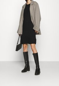 Karierte oversized Blazer, schwarzes Kleid, hohe schwarze Stiefel und eine gesteppte schwarze Handtasche. Das Outfit zeichnet sich durch kontrastierende Texturen und Muster aus.
