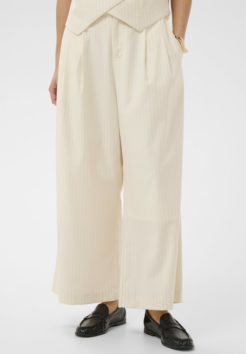 Femme portant un pantalon à larges jambes crème à rayures fines taille haute avec poches et mocassins en cuir noir, debout devant un fond uni.