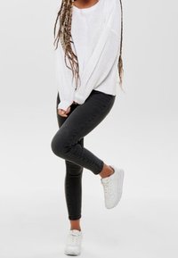 Pull en tricot blanc à manches longues, associé à un jean skinny noir et des baskets blanches, sur un fond gris uni.