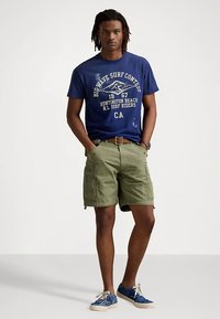 Manns avslappede antrekk med en blå T-skjorte med hvit tekst, olivengrønne cargoshorts og blå sneakers med hvite lisser.