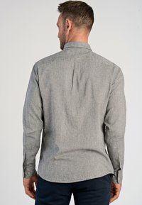 Camicia grigia a maniche lunghe con tessuto testurizzato, polsini bottonati e colletto piccolo. Presenta una piega centrale sul retro e un orlo arrotondato.