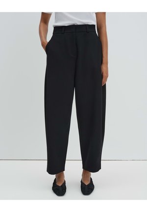 Person trägt schwarze, locker sitzende High-Waist-Hosen mit Seitentaschen, kombiniert mit schwarzen, gerafften Ballerinas und einem in die Hose gesteckten weißen Oberteil.