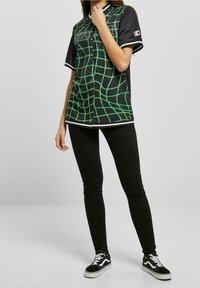 Maglietta nera in stile baseball con motivo a griglia verde, maniche corte e chiusura a snap. Abbinata a leggings neri e sneakers nere.