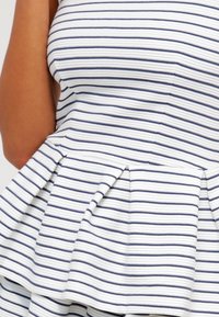 Peplum top vyrobený z bílé látky s modrými vodorovnými pruhy, s přiléhavým živůtkem a rozšířeným peplum lemem.