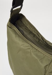 Olivnozeleni najlonasti nahrbtnik z teksturirano površino, z zgornjim zadrgo in črnimi kovinskimi dodatki. Notranjost je obložena s temnejšo tkanino.