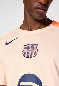 Nike shirt in licht perzik met een textuurstof, met een donkerblauwe geborduurde FC Barcelona-embleem en een blauw logo accent aan de voorkant.