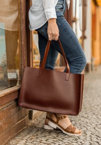 Sac cabas en cuir marron avec deux poignées robustes, forme rectangulaire, texture lisse et un logo subtil gaufré à l'avant.