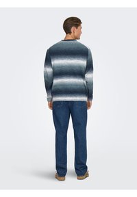 Homme debout de dos portant un pull à rayures bleu et blanc, un jean bleu et des chaussures à lacets de couleur beige, devant un fond uni.