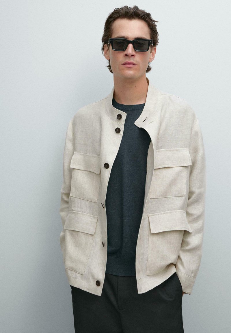 Giacca in lino beige con quattro tasche frontali, collo rotondo e bottoni marroni, indossata sopra un maglione scuro e pantaloni neri. Occhiali da sole neri.