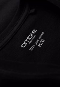 Étiquette d'un t-shirt en coton noir avec le logo "ombre", indiquant "100% coton pur" et la taille "M" en texte blanc sur un fond texturé.