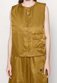 Sleeveless golden-brown zip-up vest made of shiny fabric, featuring a front pocket and subtle branding detail. Textured finish, minimal design.
