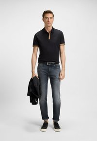 Camisa polo negra con detalles en beige, combinada con jeans oscuros y zapatillas negras. Se sostiene un blazer en la mano izquierda, con un diseño ajustado.