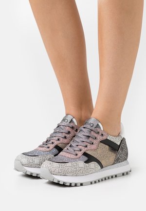 Baskets usées avec un motif multicolore, des accents scintillants argentés et roses, une bordure noire et des semelles texturées, portées sur des jambes nues sur un fond blanc.