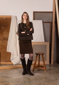 Hnedý pletený top so zlatými gombíkmi, hnedá ceruzková sukňa v rovnakom odtieni a hnedé semišové čižmy po kolená. Modelka stojí v štúdiovom prostredí.