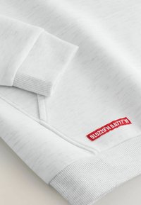 Sweatshirt blanc avec des poignets et un ourlet côtelés. Présente une étiquette rouge avec des coordonnées brodées sur le côté inférieur. Tissu doux et texturé.