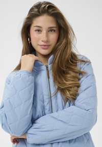 Svetlo modra prešita jakna z zadrgo spredaj, rebrastim ovratnikom in teksturirano tkanino. Modelka pozira z rokami prekrižanimi, kar poudarja dizajn.