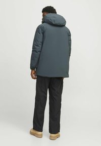 Padded parka u tamno sivoj boji s kapuljačom, ravnog kroja i bez vidljivih metalnih detalja, nosi se preko crnih cargo hlača i smeškastih čizama.