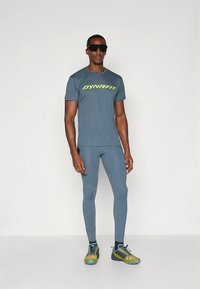 Moška športna oprava s temno modro majico s kratkimi rokavi, na kateri je napis "DYNAFIT" v svetlo rumeni barvi, v kombinaciji z usklajenimi oprijetimi legicami in tekaškimi čevlji.