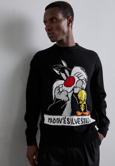 Pull en tricot noir avec un design de cartoon représentant un personnage tenant un objet rouge et un petit oiseau jaune. Le texte en dessous dit "MADOVÉŠILVESTR."