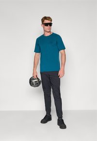 T-shirt de sport teal à manches courtes, pantalons noirs et baskets foncées. La personne tient un casque noir et porte des lunettes de soleil sombres.