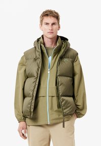 Chaqueta tipo puff en nylon verde oliva, con capucha, cierre frontal de cremallera con detalles en azul claro y puños elásticos. Diseño en capas con una textura suave.