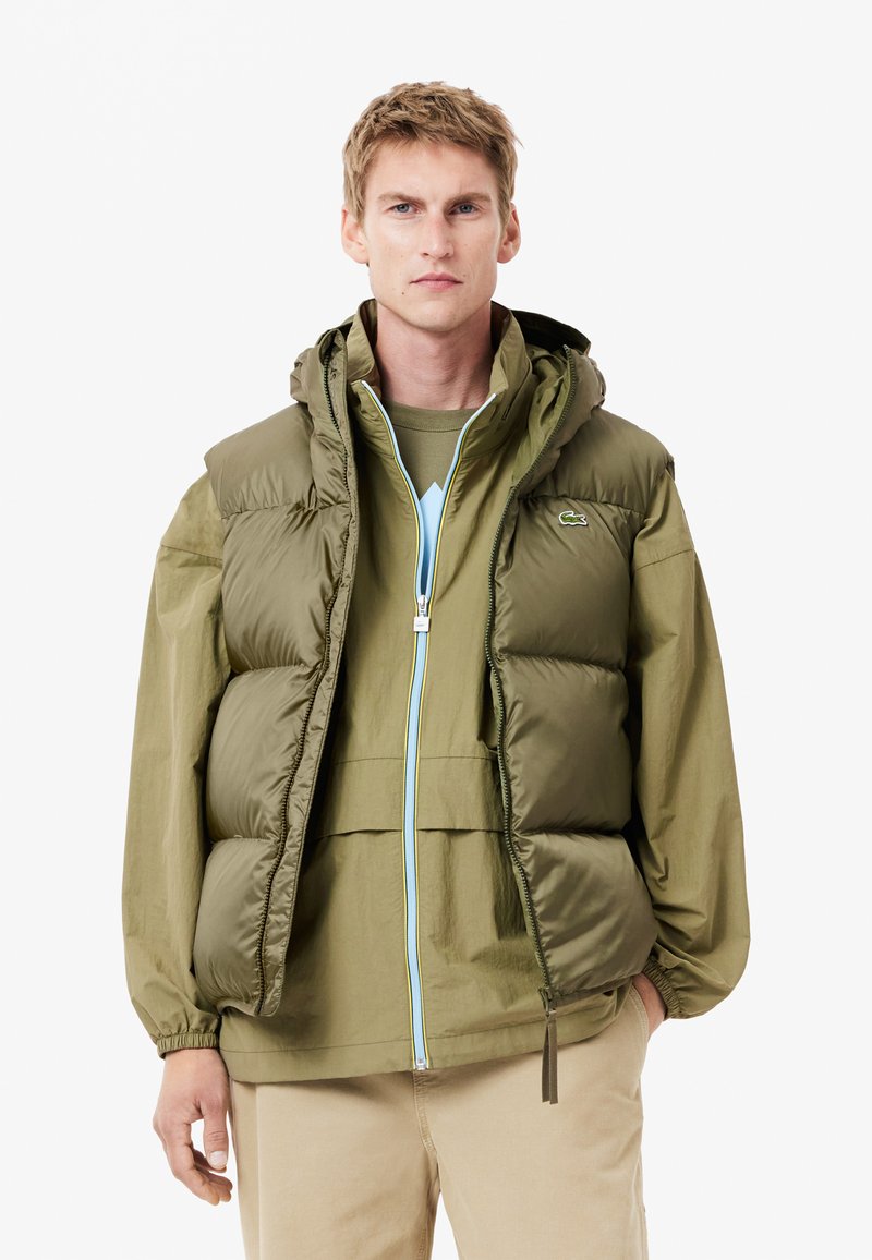 Chaqueta tipo puff en nylon verde oliva, con capucha, cierre frontal de cremallera con detalles en azul claro y puños elásticos. Diseño en capas con una textura suave.
