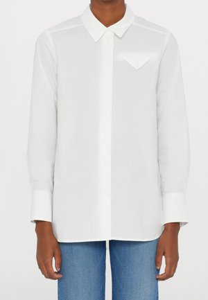 Persoon draagt een wit lang mouwenshirt met knopen en een driehoekige klep op de borstzak en blauwe spijkerbroek, staand tegen een effen achtergrond.