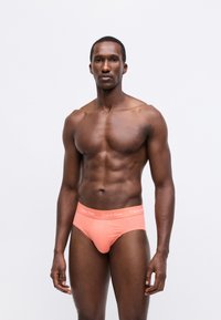 Barackszínű Calvin Klein alsónadrág, logós derékpánttal, sima anyagból, testhezálló szabással és alacsony derékmegoldással. A modellen ing nélkül látható a szabás.