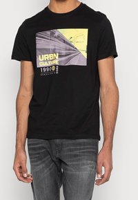 Svart bomull t-shirt med grafisk tryck i gult och grått, som föreställer urbana tågräls och texten "URBN CULTURE 1990 CORE."