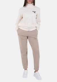 Giacca beige chiaro con cerniera e collo alto, con logo nero; abbinata a pantaloni larghi color marrone chiaro e scarpe da ginnastica bianche.