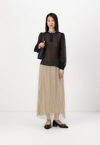 Top trasparente nero a maniche lunghe con gonna plissettata beige. Tessuto strutturato e scollatura rotonda con decorazioni. Scarpe nere con tacco e borsa.