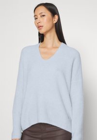 Hellblauer V-Ausschnitt-Pullover aus weichem Strickmaterial mit lockerer Passform und tiefen Schultern, der einen leicht abgerundeten Saum aufweist.