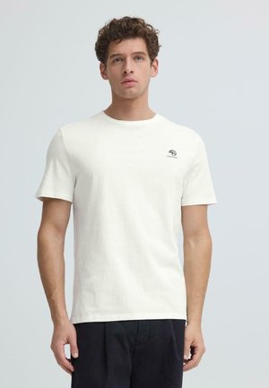 Nuori mies kiharatukkaisena, yllään yksinkertainen valkoinen lyhythihainen t-paita ja tummat housut, seisomassa vaaleanharmaan taustan edessä.