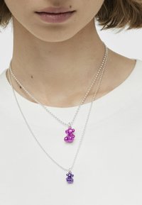 Dos collares de cuentas con colgantes de ositos de peluche metálicos, uno púrpura y otro púrpura oscuro, sobre una cadena de bolas plateada, llevados sobre una blusa blanca.