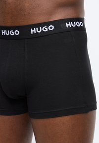 Μαύρα ανδρικά μπόξερ με φαρδύ ελαστικό περίμετρο διαθέτοντας το λογότυπο "HUGO" σε λευκά γράμματα, φορεμένα από άτομο με σκούρο χρώμα δέρματος.