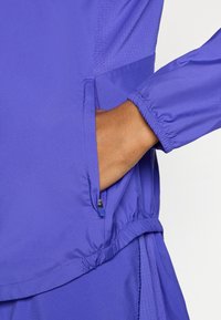 Μοβ ελαφρύ μπουφάν με φερμουάρ σε πλαϊνή τσέπη, ελαστικά μανίκια και υφή στο σώμα για αναπνοή. Λείο φινίρισμα υφάσματος.