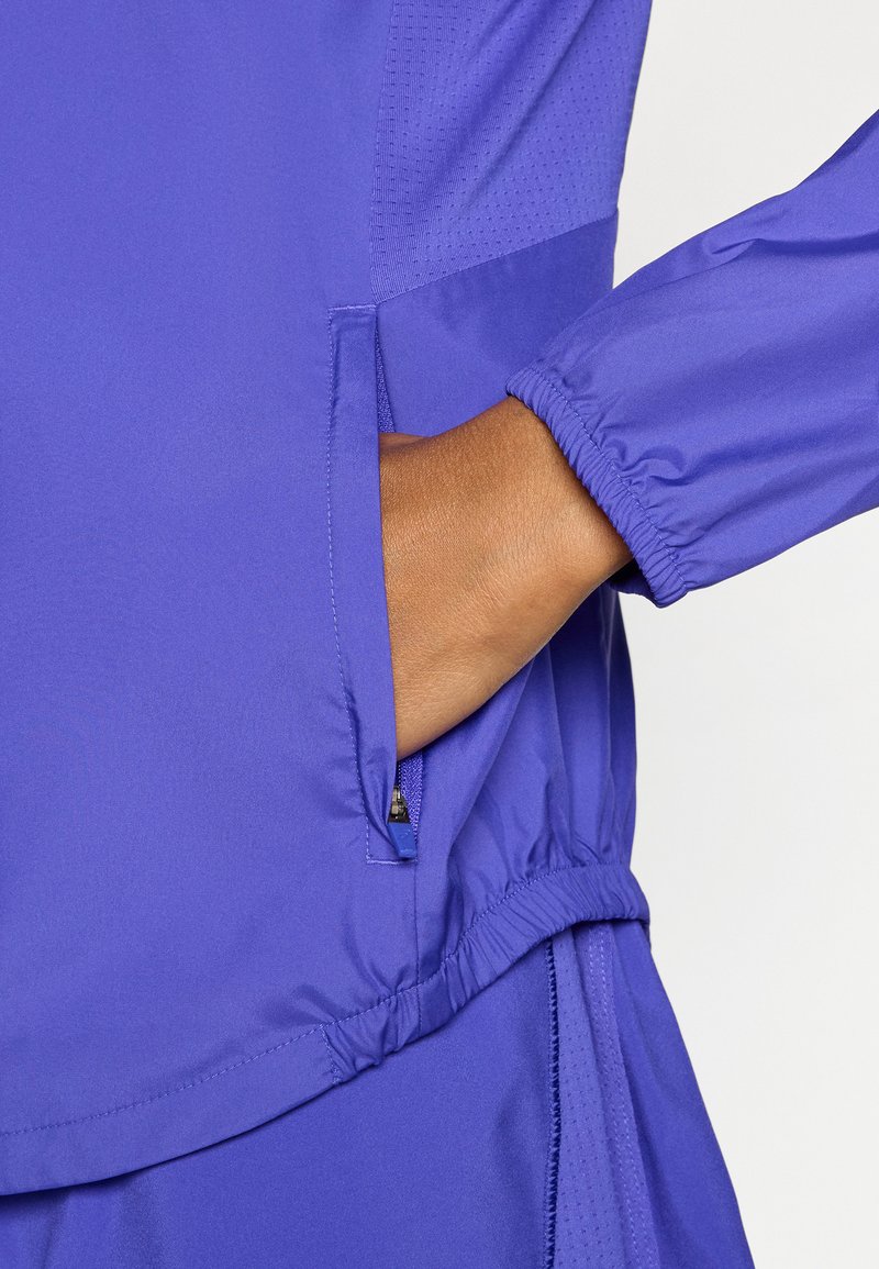 Veste légère violette avec une poche latérale zippée, des manches élastiquées et un corps texturé pour la respirabilité. Finition en tissu lisse.