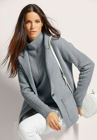 Grijze gebreide cardigan met een geribbelde textuur, gedragen over een grijze coltrui. Witte broek en een witte schoudertas met een cirkelvormig ontwerp maken de outfit compleet.