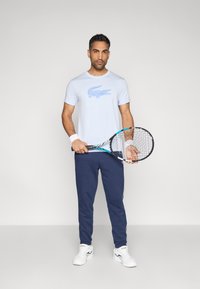 Gaiši zils īsroku t-krekls ar krokodila logo, kopā ar tumši zilām sporta biksēm. Modelis tur rokā tenisa raketi un valkā baltas kedas.