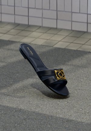 Sandale à glissière noire Aldo avec double lanières et une boucle décorative dorée sur une surface en asphalte près d'un pavage carrelé.