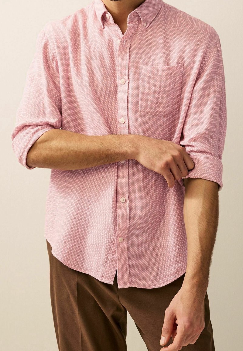 Homme remontant la manche d'une chemise rose clair à boutons, portant un pantalon marron sur un fond uni.