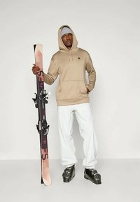 Μπεζ φούτερ με λογότυπο, λευκά παντελόνια και μαύρες μπότες σκι. Κρατώντας ένα ροζ και μαύρο σκι. Απλός σχεδιασμός, άνετη εφαρμογή και χαλαρή υφή.