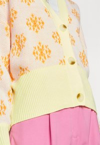 Cardigan en tricot avec un motif de flocon de neige orange sur un fond rose pâle, présentant un ourlet côtelé jaune et de grands boutons beiges.