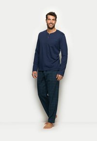 Dunkelblaues Langarm-Pyjama-Oberteil mit Knopfleiste, kombiniert mit grün-blauen karrierten Hosen. Glattes Material, lockere Passform, barfuß Model.