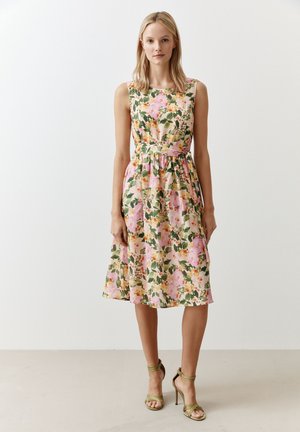 Blumiges Midi-Kleid mit tailliertem Oberteil, in einem rosa-grünen Farbschema, mit einem taillierten Bund und einem ausgestellt geschnittenen Rock. Grünes, strappy High-Heel-Sandalen.