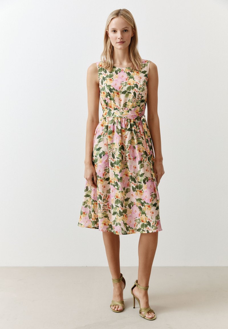 Blumiges Midi-Kleid mit tailliertem Oberteil, in einem rosa-grünen Farbschema, mit einem taillierten Bund und einem ausgestellt geschnittenen Rock. Grünes, strappy High-Heel-Sandalen.