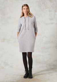 Grå luvklänning med avslappnad passform, långa ärmar, kängurufickor och justerbar dragsko. Bärs över svarta tights och stövlar.
