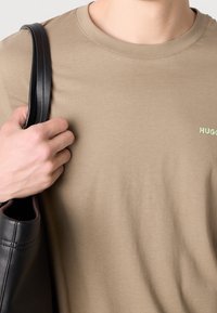 Nærbillede af person, der bærer en beige Hugo T-shirt, og holder en sort lædertaske-strop over skulderen med venstre hånd.