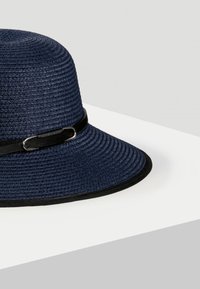 Granatowy pleciony kapelusz słomkowy z szerokim brzegiem, akcentowany czarną taśmą i srebrnymi dodatkami, charakteryzujący się teksturowaną powierzchnią i zakrzywionym kształtem.